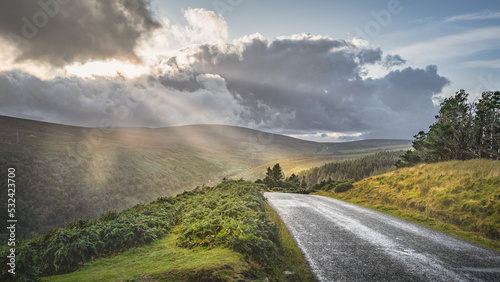 Winding road leading to Lough Tay called The Guinness Lake illuminated by sunrays from dramatic sunset sky with rain clouds Wicklow Mountains, Ireland