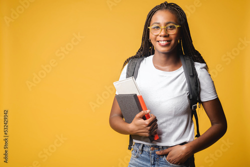 Inspired multiracial female student in white t-shirt and jeans with backpack isolated on yellow, foreigner woman in glasses proud to be university member, looking at the camera standing indoors