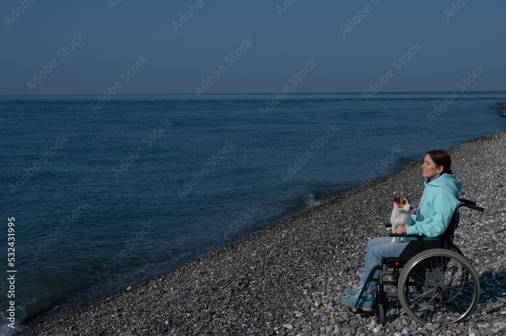 A serene Caucasian woman in a wheelchair is resting on the seashore with a jack russell terrier dog.