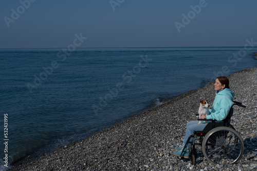A serene Caucasian woman in a wheelchair is resting on the seashore with a jack russell terrier dog.