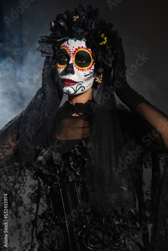 woman in sugar skull makeup and costume with black wreath and lace veil on dark background with smoke.