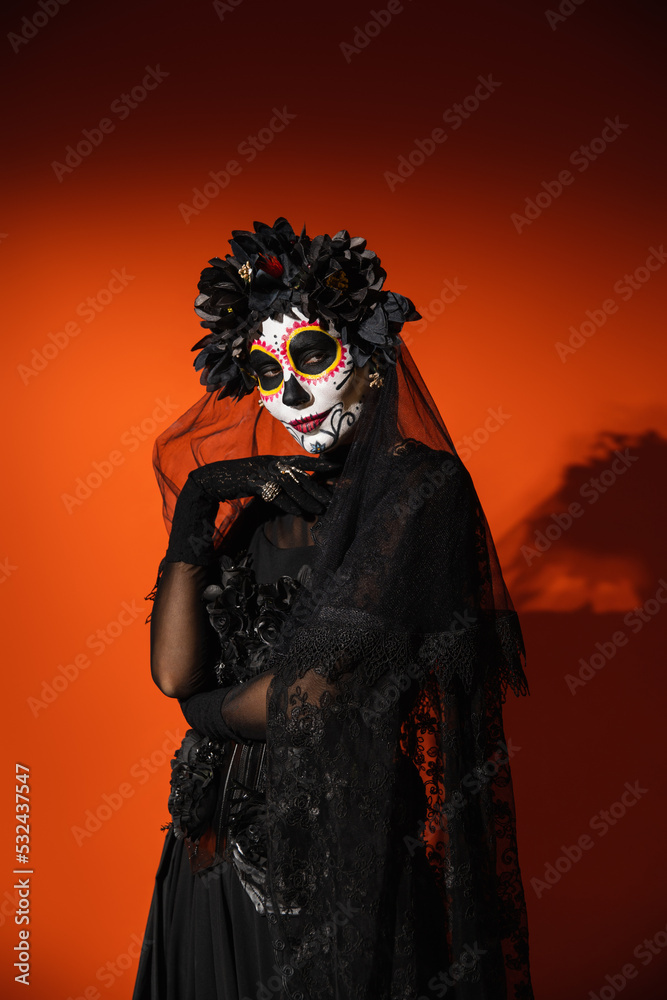 Woman in creepy mexican day of death costume and makeup looking at camera on red background.