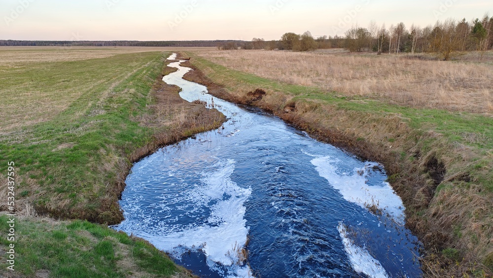 A small river runs through grassy meadows to the forest on the horizon. After passing the dam, the water in the river bubbles and foams. The spring evening twilight is setting
