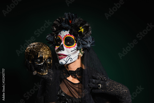 Smiling woman in santa muerte halloween costume holding skull on dark green background.