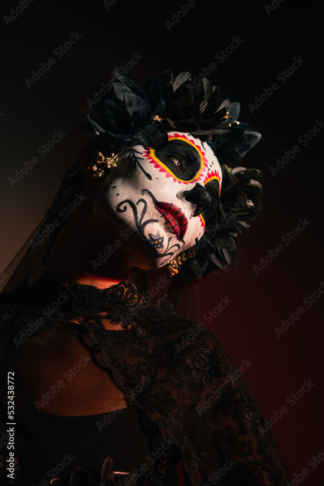 Woman in wreath with black flowers and catrina makeup looking away on burgundy background.