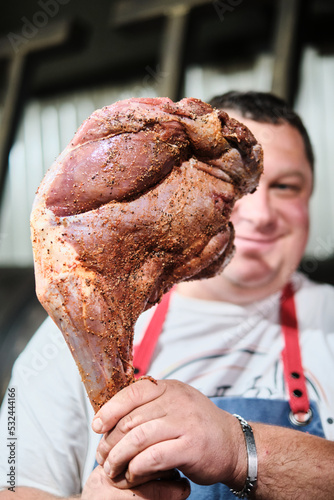 The butcher holds a leg of lamb seasoned with marinade in front of a large smoker
