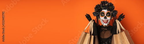 excited woman in spooky halloween makeup and black wreath holding shopping bags isolated on orange, banner.