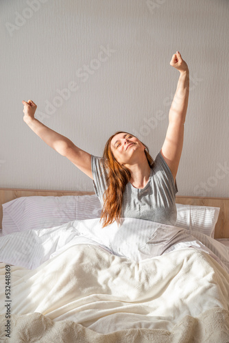 Woman just waking up and stretching in bed at home in the morning. Rested and relaxed happy Lady with long hair in pajamas. Girl on clean white bedding with cozy blanket. Healthy lifestyle, wellness photo