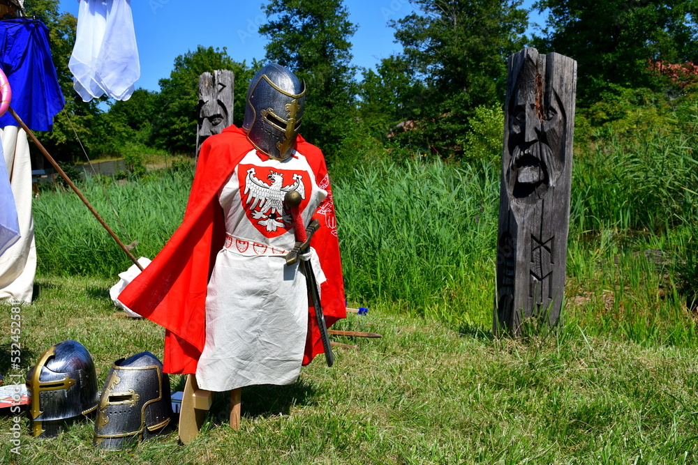 A close up on a set of medieval knight armor with a Polish crest on the chestpiece seen next to some helmets and nordic figurines in the middle of a public park during a medieval fair or festival