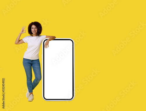 Gesturing OK young african american woman happy leaned on big, huge smartphone with white screen wearing white t-shirt and denim jeans isolated on yellow background. App advertisement mock up