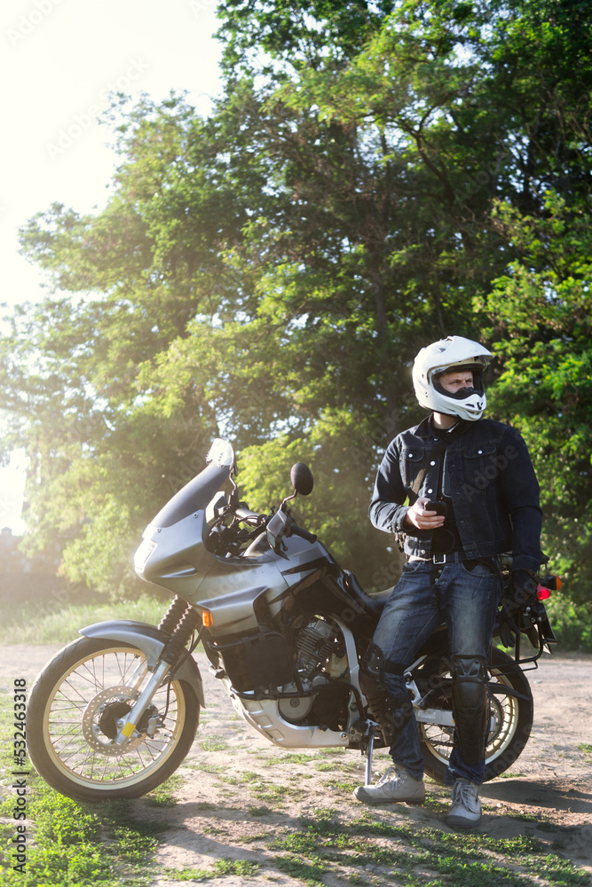 Fototapeta premium rider guy use smart phone in jeans biker jacket and helmet sit on tourist touring motorcycle. outdoors, dual sport adventure concept, vertical photo, dirt road