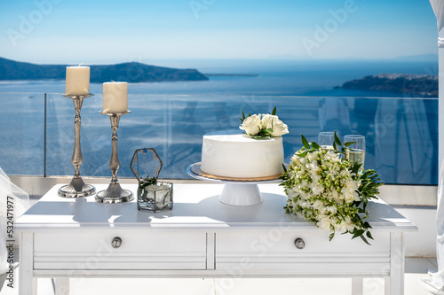 Fototapeta Naklejka Na Ścianę i Meble -  Bridal bouquet made of white freesia flowers, wedding cake, two glasses with sparkling wine, two candlestick on the white table. Blurred blue sea at the background.  Wedding details. 