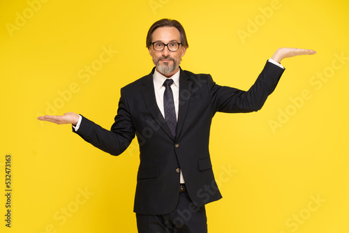 Leveling up, weighing or balancing middle aged business man with hands up in levels looking at camera isolated on yellow background. Handsome mature businessman in black suit. Business concept