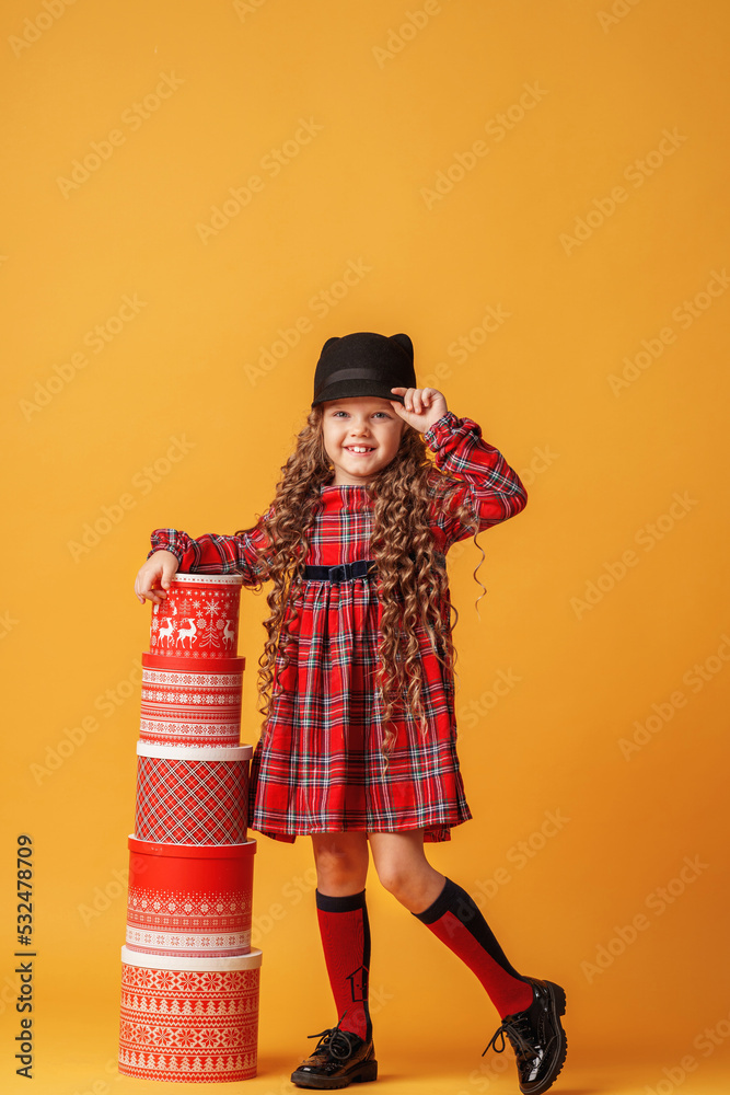 little girl in hat and plaid dress plays with Christmas gift boxes, building pyramid on yellow background in studio. the child is happy to receive many gifts for Christmas. Advertising and discounts. little girl in hat and plaid dress plays with Christmas gift boxes, building pyramid on yellow background in studio. the child is happy to receive many gifts for Christmas. Advertising and discounts.