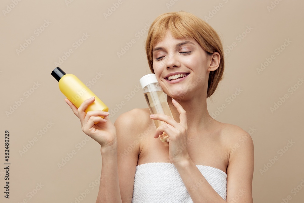 Fototapeta premium a happy, friendly woman with clean skin, stands on a beige background and advertises jars of cosmetics in her hands, smiling with her eyes closed, lifting them to her face.
