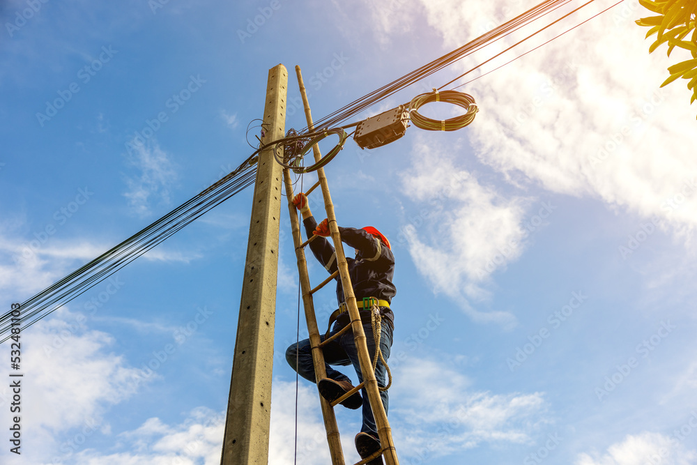 A telecoms worker is shown working from a utility pole ladder while wearing high visibility personal safety clothing, PPE, and a hard hat.. - obrazy, fototapety, plakaty 