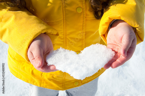 On a bright sunny day in winter, a little girl in a yellow jacket holds a heart made of snow in her hands.