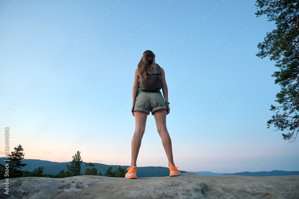 Sportive woman standing alone on hillside trail. Female hiker enjoying view of evening nature from rocky cliff on wilderness path. Active lifestyle concept