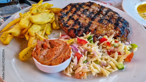 Greek burgers (biftekia) served with red sauce, fried potatoes and salad in an authentic restaurant in streets of Thessaloniki, Central Macedonia, Greece, Europe. Mince meat beef patty on white plate