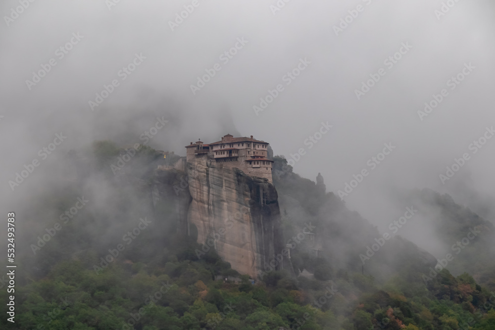 Fototapeta premium Panoramic view of Holy Orthodox Monastery of Rousanou (St. Barbara), Kalambaka, Meteora, Thessaly, Greece, Europe. Unique rock formation are surrounded by mystical fog. Moody dramatic atmosphere
