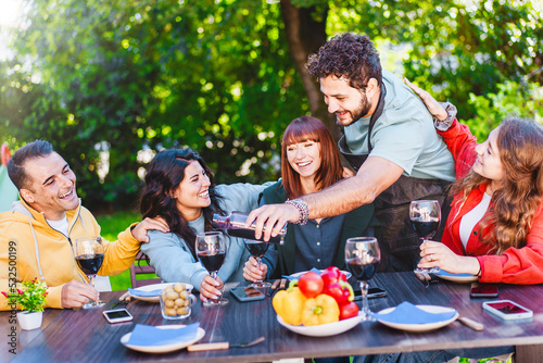 Man pouring red wine into glass while enjoying dinner party with friends outdoors in the backyard