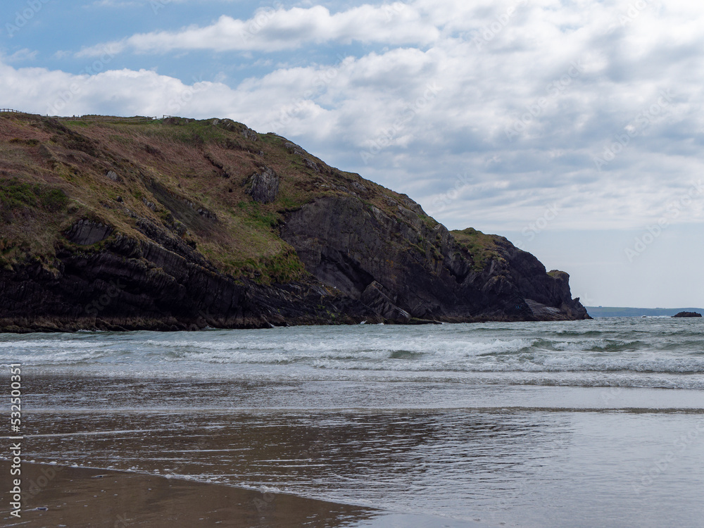 View of the coastal cliffs from the Warren beach. The rocky coast of the south of Ireland. Hills under the sky on a spring day, mountain beside sea, clouds. View of the coastal cliffs from the Warren beach. The rocky coast of the south of Ireland. Hills under the sky on a spring day, mountain beside sea, clouds.