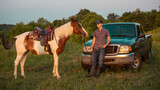 A cowboy on a ranch near a pickup truck. Western horse on the field at sunset. Cowboy saddle and boots.