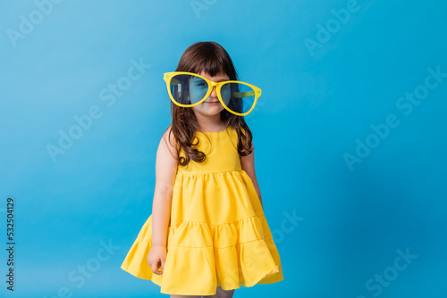 Bilde på lerret cute little girl in a yellow dress and huge sunglasses stands on a blue background