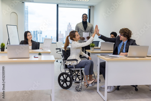 Handicap business woman giving high-five to colleague in creative office. Office workers and woman in a wheelchair are making a conversation in bright office. They are showing a teamwork.