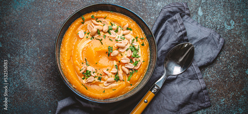 A ceramic bowl with pumpkin cream soup decorated with peanuts on dark stone background with spoon decorated, from above. Autumn cozy dinner concept 
