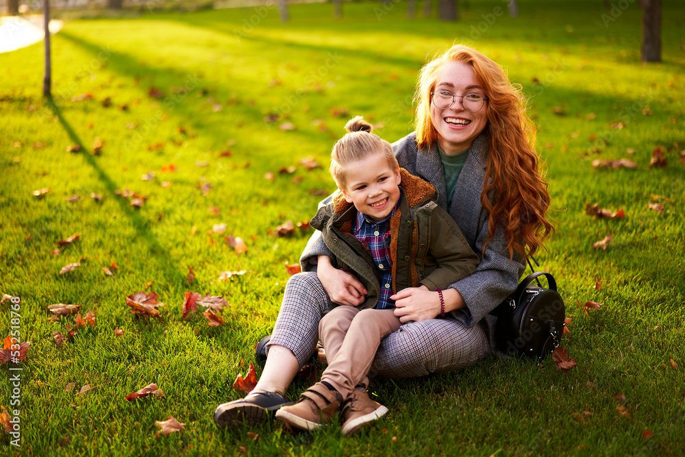Smiling redhead woman and little boy sitting on grass lawn in city park on warm autumn day. Young mom hugs her son, they have fun and look at camera on sunny fall day. Foliage on green lawn. Smiling redhead woman and little boy sitting on grass lawn in city park on warm autumn day. Young mom hugs her son, they have fun and look at camera on sunny fall day. Foliage on green lawn.