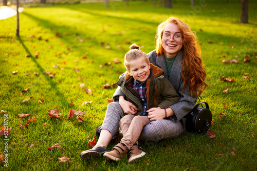 Smiling redhead woman and little boy sitting on grass lawn in city park on warm autumn day. Young mom hugs her son, they have fun and look at camera on sunny fall day. Foliage on green lawn.