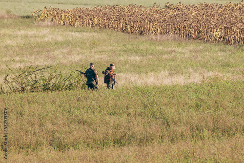 Two hunters armed with rifles wait to shoot their prey in a field with dried sunflowers in the background