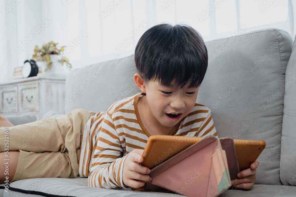 Handsome Asian boy, lying on sofa in his comfortable position, along with hands on feet, chin, Boy laying down watching online clips on his tablet merrily, his face looked happy.