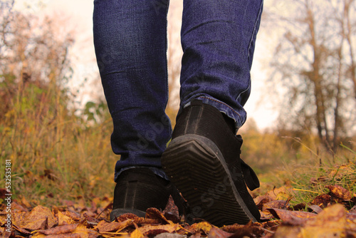 Women's legs in blue jeans and black sneakers against the background of autumn yellow-orange leaves. Back view