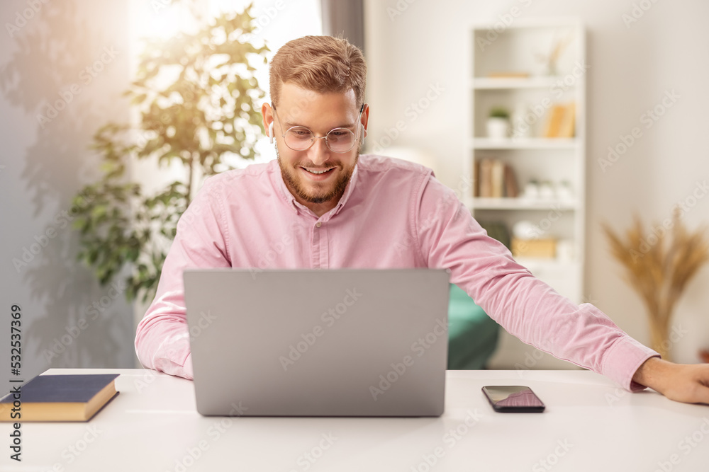 Handsome bearded man in eyewear working on laptop, happy male work from home, light interior of living room Handsome bearded man in eyewear working on laptop, happy male work from home, light interior of living room