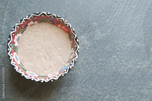 Top view of dry clay (rhassoul, ghassoul) in a colorful porcelain bowl on a gray stone background.