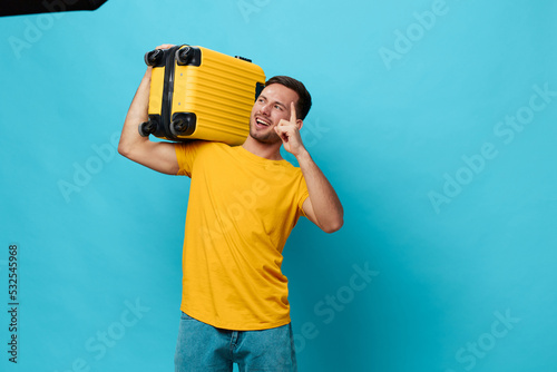 Overjoyed tanned handsome man in yellow t-shirt point finger up ready to vacation look aside hold suitcase posing isolated on blue studio background. Copy space Banner Mockup. Trip journeys concept