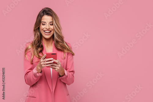 Photo of a happy young beautiful woman using mobile phone, smiling, posing isolated over pink pastel background.
