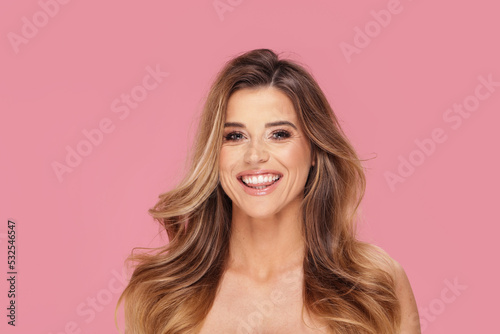 Happy young beautiful woman in glamour makeup and long wavy hair smiling to the camera  posing isolated on pink pastel studio background. Beauty portrait.