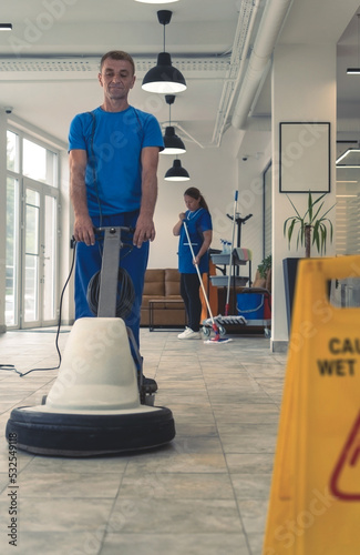 Senior cleaner cleans the hard floor with machine while cleaning lady cleans in the backround