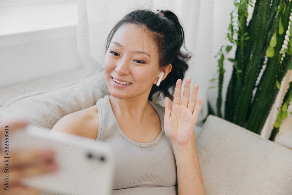Athletic Asian woman in yoga costume blogger filming herself on phone camera sitting on couch and smiling with headphones waving, freelance work from home, recording sports workout lifestyle Athletic Asian woman in yoga costume blogger filming herself on phone camera sitting on couch and smiling with headphones waving, freelance work from home, recording sports workout lifestyle