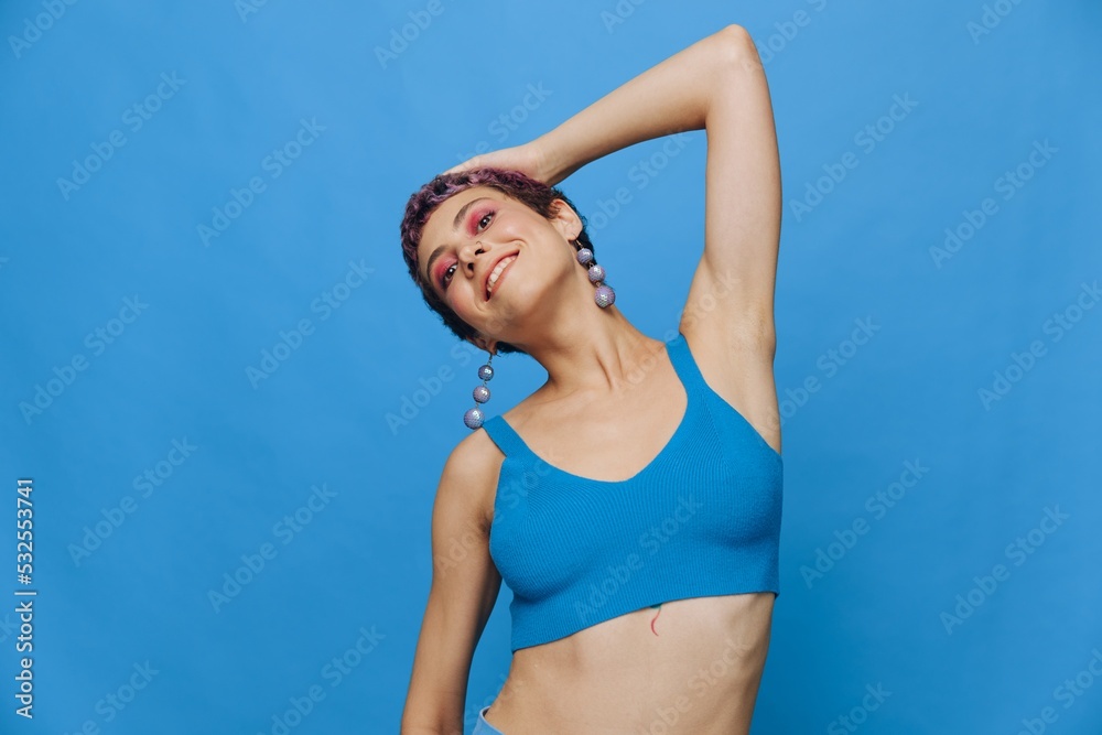 Fototapeta premium Young athletic fashion woman with colored hair and short haircut posing and dancing in blue sportswear smiling and looking at the camera on a blue monochrome background