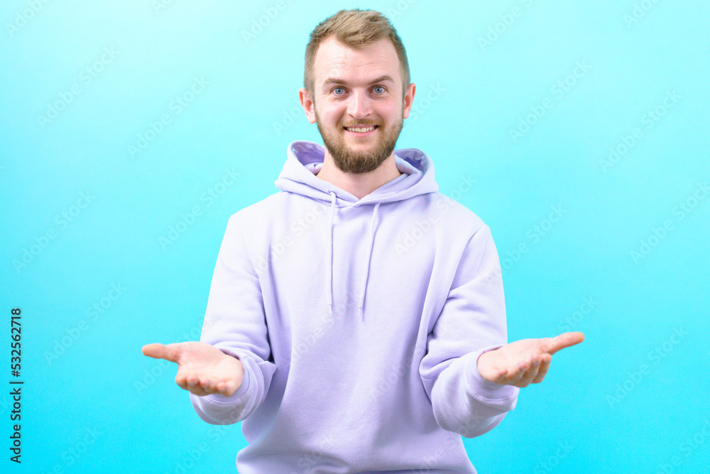 Man in a purple hoodie hold some object and smiling at the camera on a blue background. Presenting. Offering. Blank. Portrait. Guy. Handsome. Hope. Positive. Accessibility. Background. Young Man in a purple hoodie hold some object and smiling at the camera on a blue background. Presenting. Offering. Blank. Portrait. Guy. Handsome. Hope. Positive. Accessibility. Background. Young