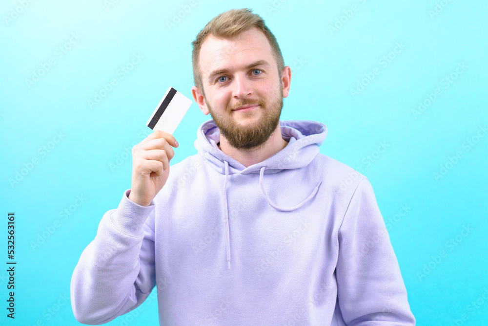 A smiling young man in a casual purple hoodie is holding a bank card on a blue background. Commerce. Studio. Bill. Blue. Beauty. Attractive. Bonus. Cashless. Buying. Lifestyle. Handsome. Commercial A smiling young man in a casual purple hoodie is holding a bank card on a blue background. Commerce. Studio. Bill. Blue. Beauty. Attractive. Bonus. Cashless. Buying. Lifestyle. Handsome. Commercial