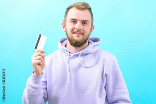 A smiling young man in a casual purple hoodie is holding a bank card on a blue background. Young. Card. Online. Pay. Payment. Boy. Looking. Business. Modern. Banking. Currency. Purchase. Rich. Blond