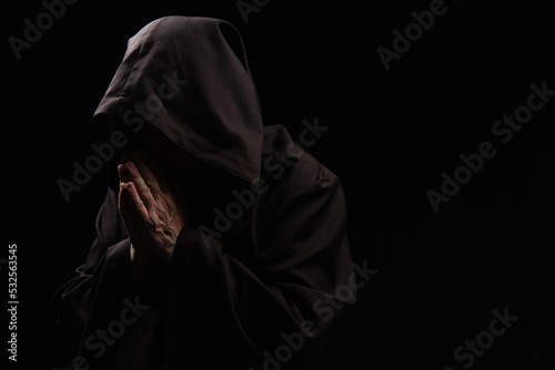 mysterious priest in hooded robe praying isolated on black.