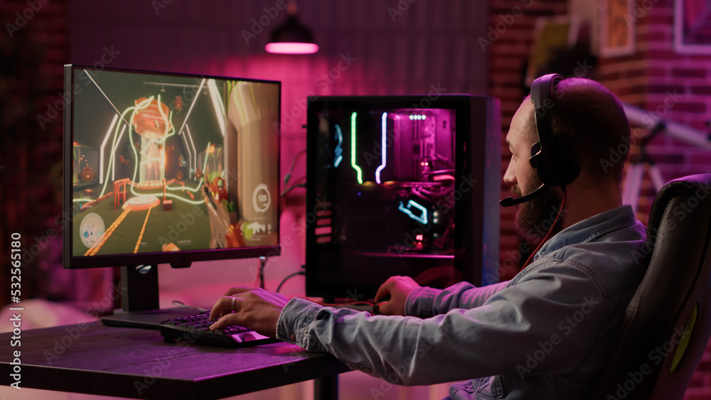 Static tripod shot of man using pc setup playing multiplayer online action game talking to team on headset. Gamer sitting in gaming chair streaming first person shooter while explaining gameplay.