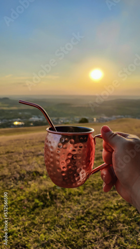 Mann hält Cocktailbecher auf einer Wiese Richtung Sonnenuntergang photo