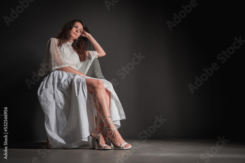 Photo of young, charming, pensive, positive pretty girl, hands, cheeks, sit on chair, wearing light dress, isolated on gray background